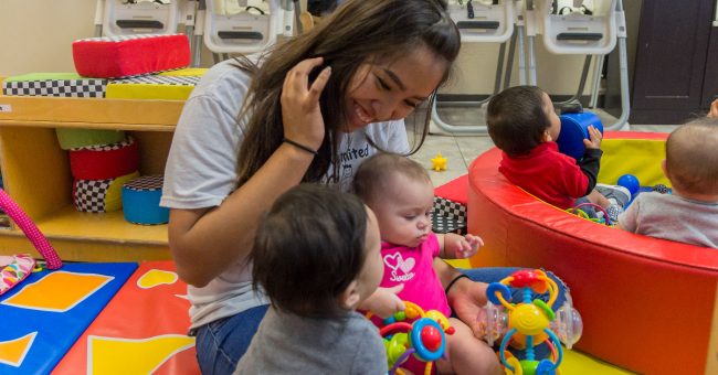 child care provider holding baby in pink in a classroom. Small boy in the fore ground.