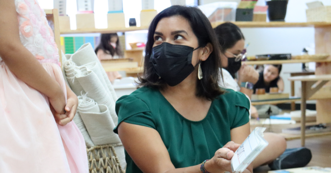 Teacher, wearing a mask, is looking up to student, pointing at a card. Quality First program.